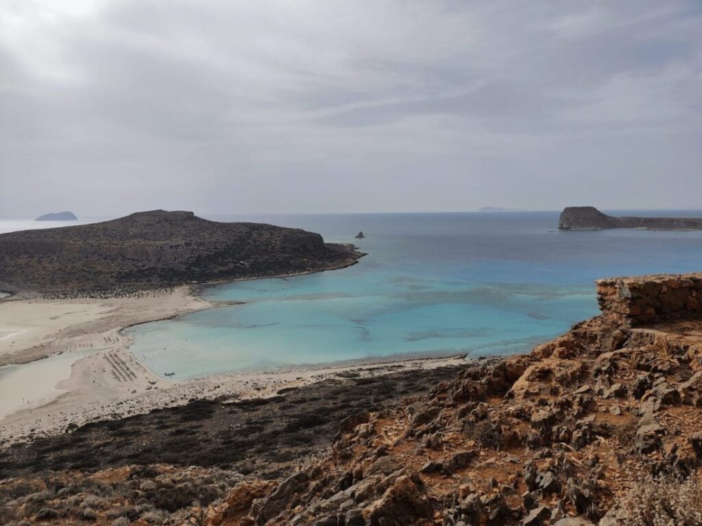 A panoramic view of Balos Lagoon in Crete, showcasing its stunning turquoise waters, sandy beach, and surrounding rugged cliffs under a slightly overcast sky.
