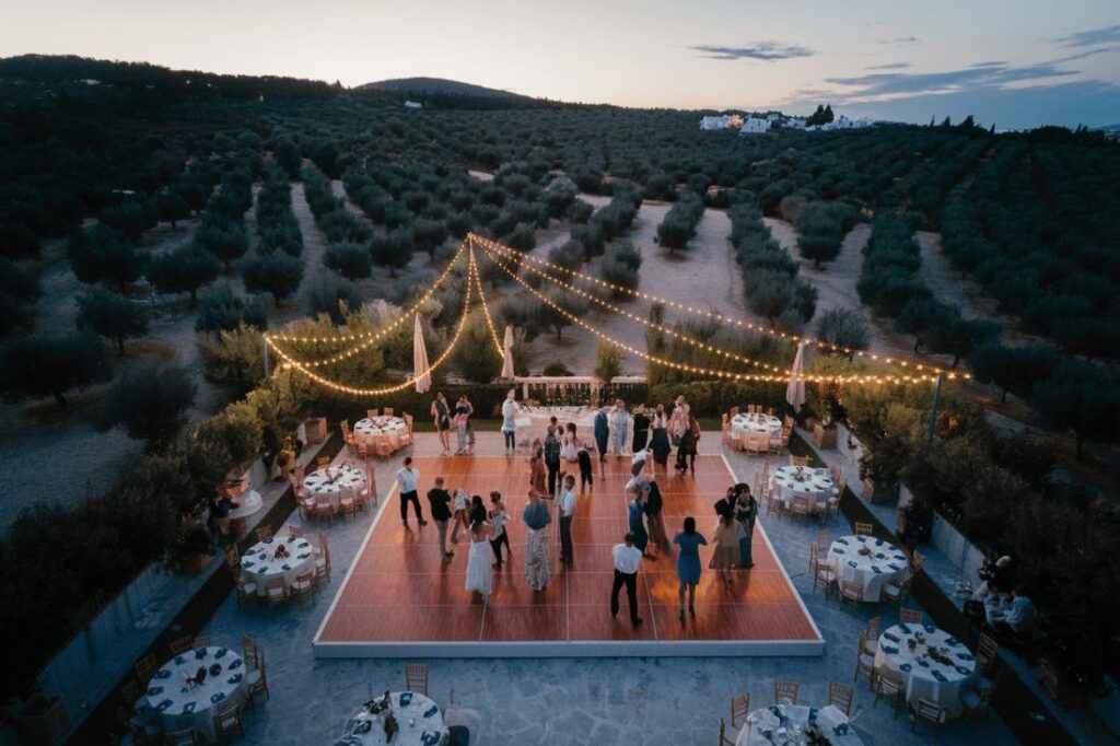 Aerial view of a wedding reception in Greece featuring a dance floor and elegantly set tables surrounded by olive trees and twinkling lights at sunset.