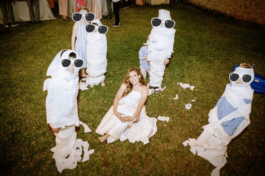 A group of children dressed in white, resembling mummies, are playfully wrapped in toilet paper around a woman in a wedding dress sitting on the grass. The children are wearing sunglasses and appear to be having fun.
