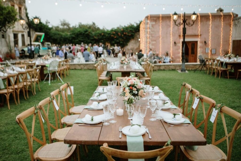 A beautifully set outdoor dining area for a wedding, featuring wooden tables adorned with flower centerpieces and elegant tableware, surrounded by guests enjoying the celebration.