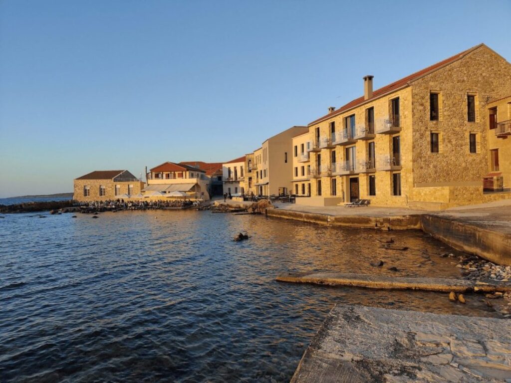 A picturesque view of a coastal area in Chania, Greece featuring charming stone buildings, local cafes, and calm waters under a clear blue sky.