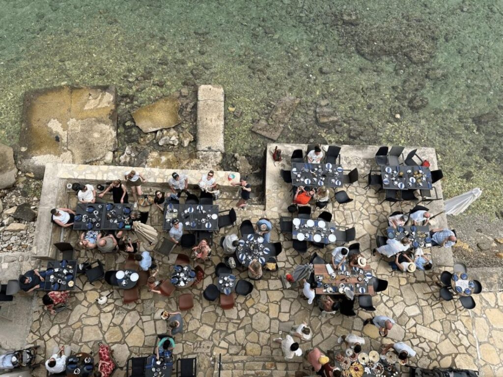 An aerial view of a seaside restaurant with multiple outdoor tables set for dining, surrounded by clear water and rocky shoreline, showcasing guests enjoying their meals.