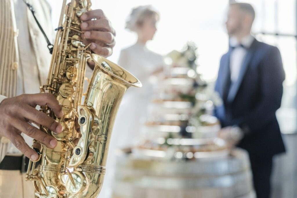 A musician playing a saxophone at a destination wedding in Greece, with a bride and groom blurred in the background near a decorative display.