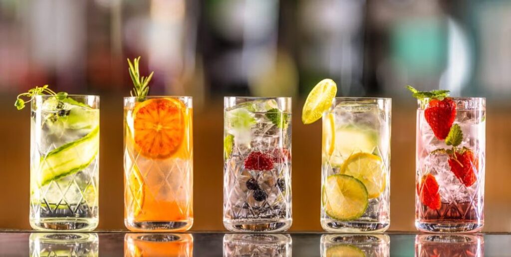 Five colorful cocktails arranged in tall glasses, featuring various fruits like cucumber, orange, lime, blueberries, and strawberries, all garnished with fresh herbs.