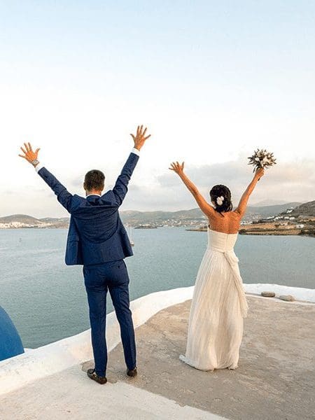 A Married Couple Getting married in Santorini, Greece, facing the sea and lifting their hands up in their air.