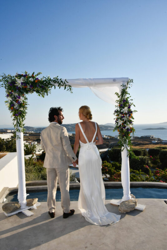 bride and groom stand in front of a wedding arch at aletro cottage houses in mykonos greece