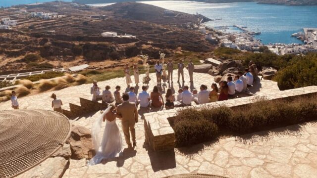 A wedding ceremony in the stone amphitheatre at Alma on Ios, guests seated on the terraces above the port and the bay.