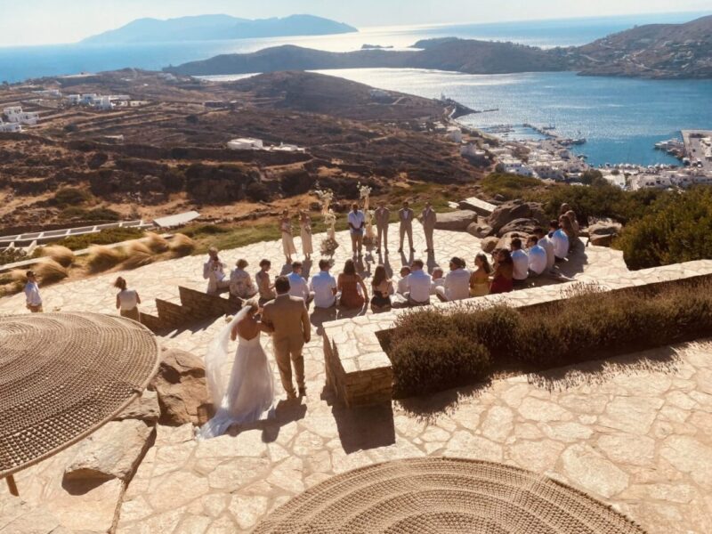 A wedding ceremony in the stone amphitheatre at Alma on Ios, guests seated on the terraces above the port and the bay.