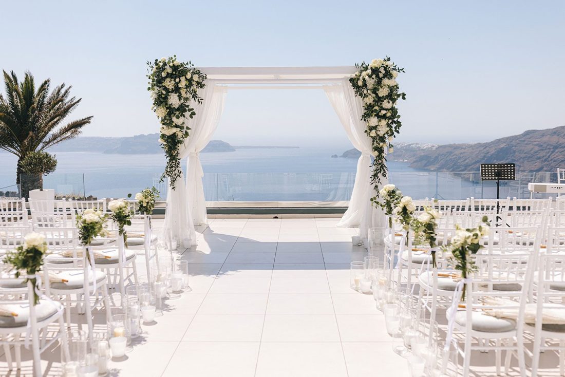 Ceremony terrace at Le Ciel Santorini with a white draped arch, rows of white chairs and the caldera sea behind