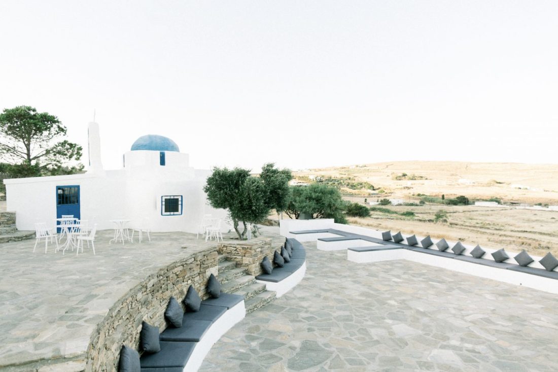 The white and blue domed chapel of St. Marina at The Secret View at Fladakia Estate, Paros, with curved stone seating terraces and an olive tree