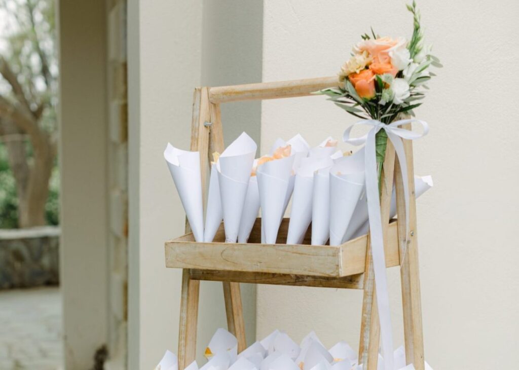 A wooden display stand holding wedding white paper cones filled with flower petals, adorned with a small bouquet of roses and greenery tied with a ribbon.