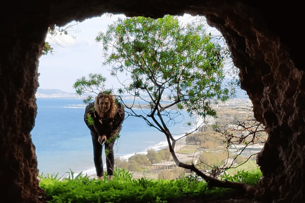 Christina bending down to examine greenery while framed by a rocky opening, revealing a stunning coastal view of the sea and beach in the background.