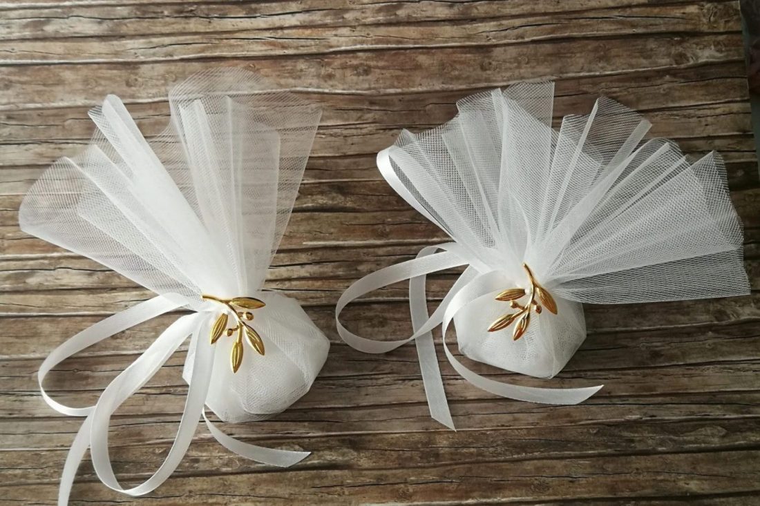 Greek wedding favors in Two white tulle pouches tied with ribbons and adorned with golden olive branch decorations, placed on a wooden surface.