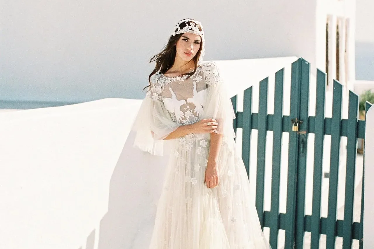 Bride in an embroidered tulle gown and lace headpiece beside a teal gate and whitewashed wall, sea behind