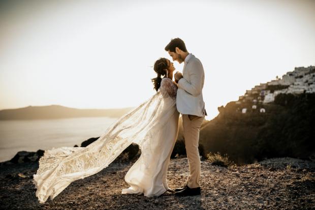 Bride and groom on a Santorini clifftop at sunset, her lace cape blowing, the caldera and village behind