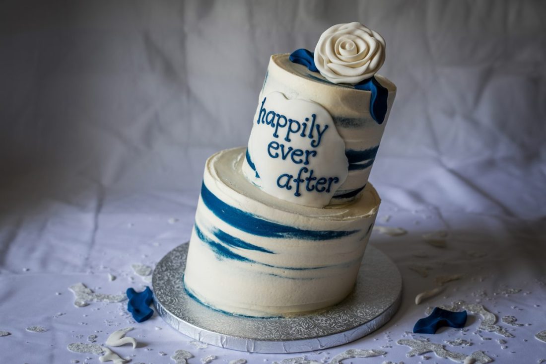 A two-tier wedding cake decorated with swirling blue and white icing, topped with a white rose and featuring the phrase 'happily ever after' written in blue icing.