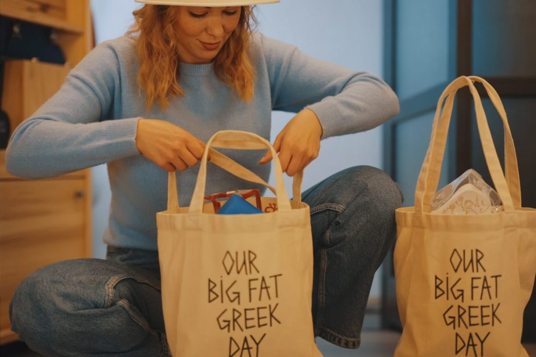 Christina, the author of Big Fat Greek Day, wearing a light blue sweater and a white hat is packing items into personalized canvas tote bags that read 'OUR BIG FAT GREEK DAY'.