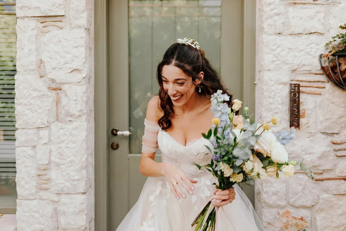 A laughing bride in the doorway of a stone house, holding a loose garden-style bouquet of pale blue, white and yellow flowers