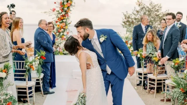 a newlywed couple takes their first kiss while walking down the aisle on a greek island