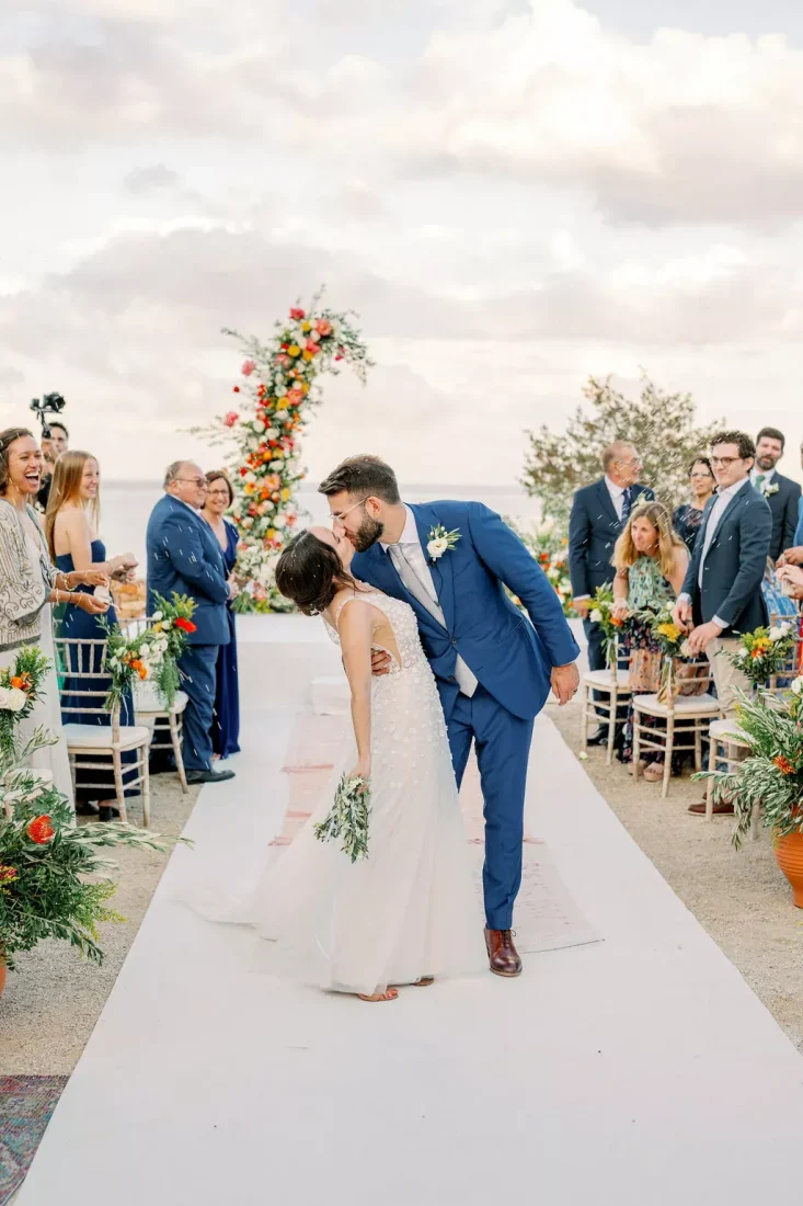 a newlywed couple takes their first kiss while walking down the aisle on a greek island
