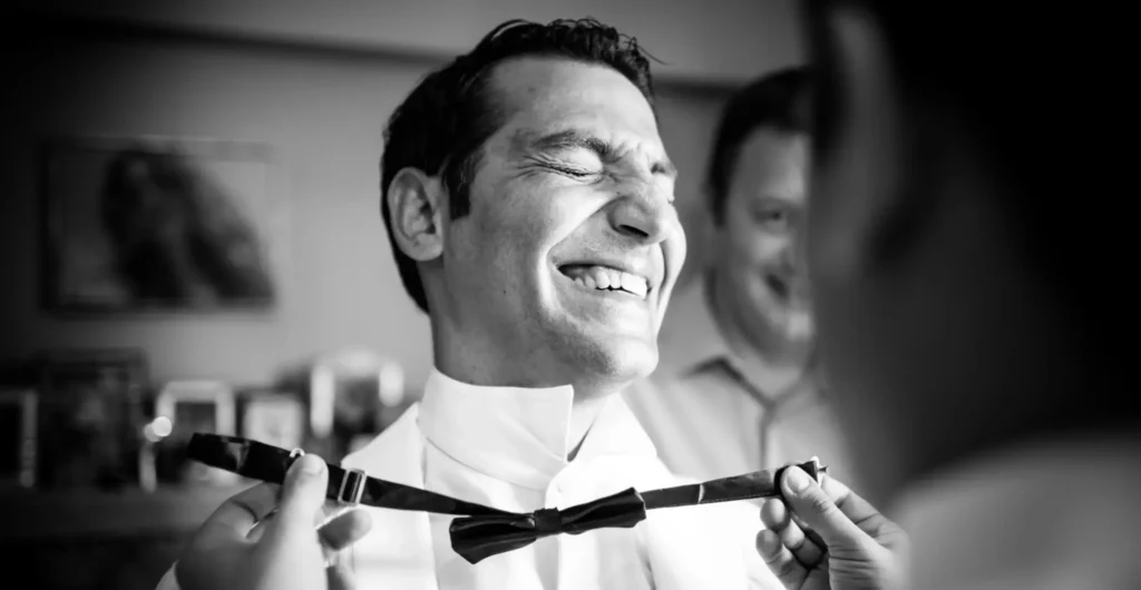 groom being dressed by his koumbaro putting on a black tie on his tuxedo