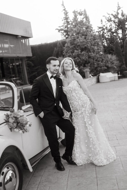 Black and white photo of a couple leaning against a vintage car decorated with flowers