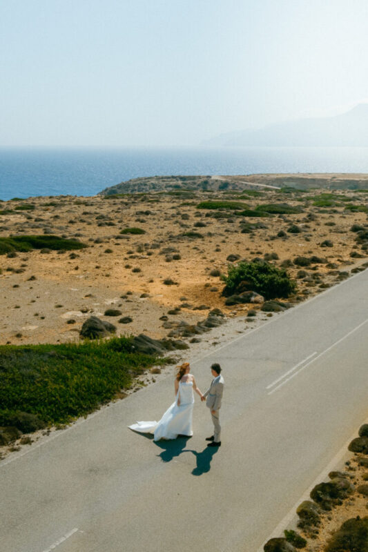 Couple holding hands on an empty coastal road, seen from above, with the sea behind them