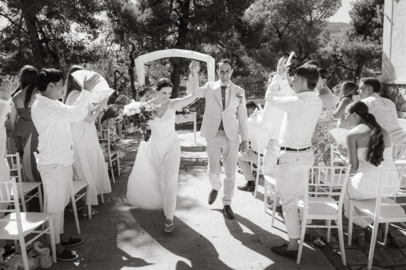Black and white photo of a bride and groom walking back down the aisle under pine trees as guests cheer