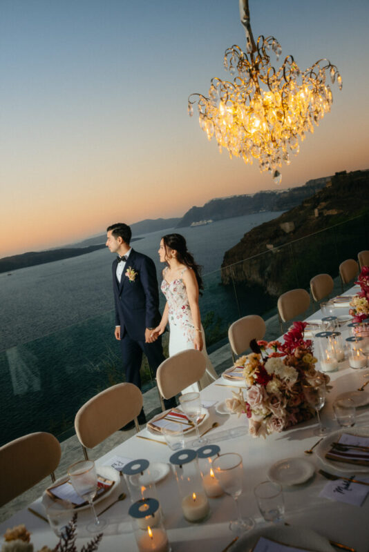 Couple beside a long candlelit dinner table under a chandelier, looking out over the sea at sunset