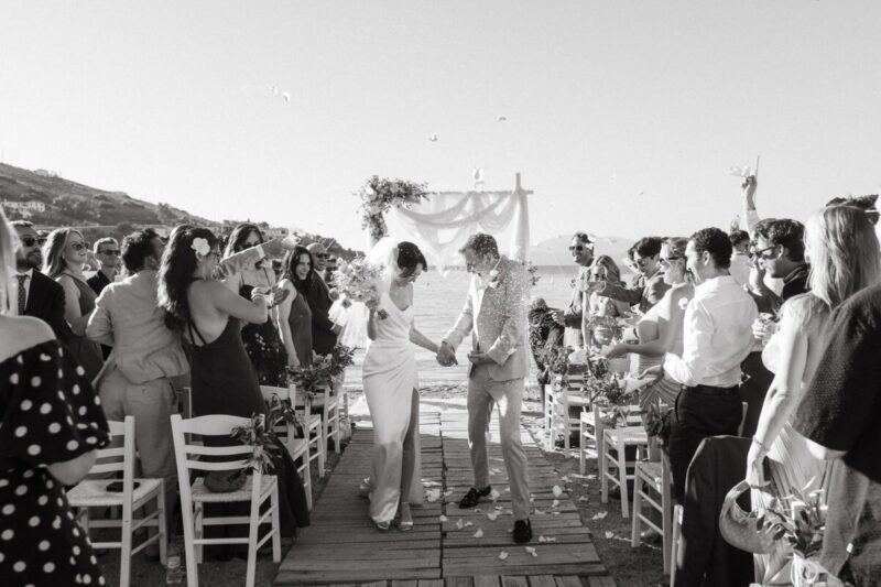 Black and white photo of a couple walking back down a wooden aisle by the sea as guests cheer