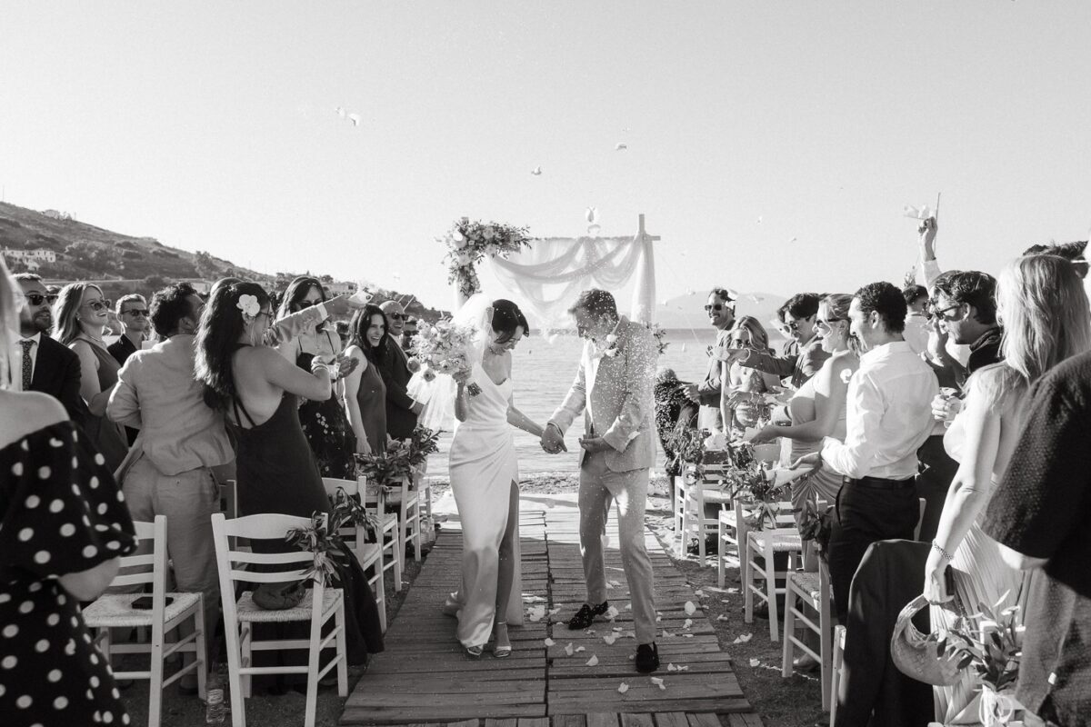 Black and white photo of a couple walking back down a wooden aisle by the sea as guests cheer