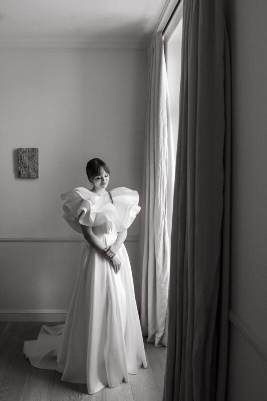 Black and white photo of a bride standing by a tall window