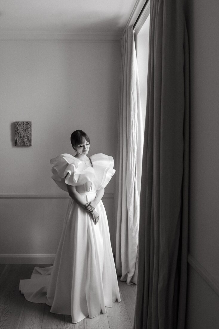 Black and white photo of a bride standing by a tall window