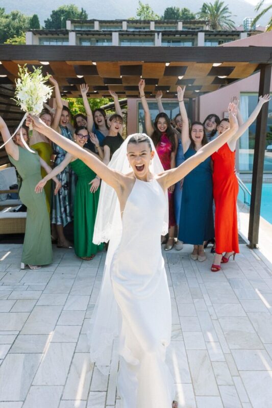 Bride with her arms raised in celebration as her bridesmaids cheer behind her by a pool