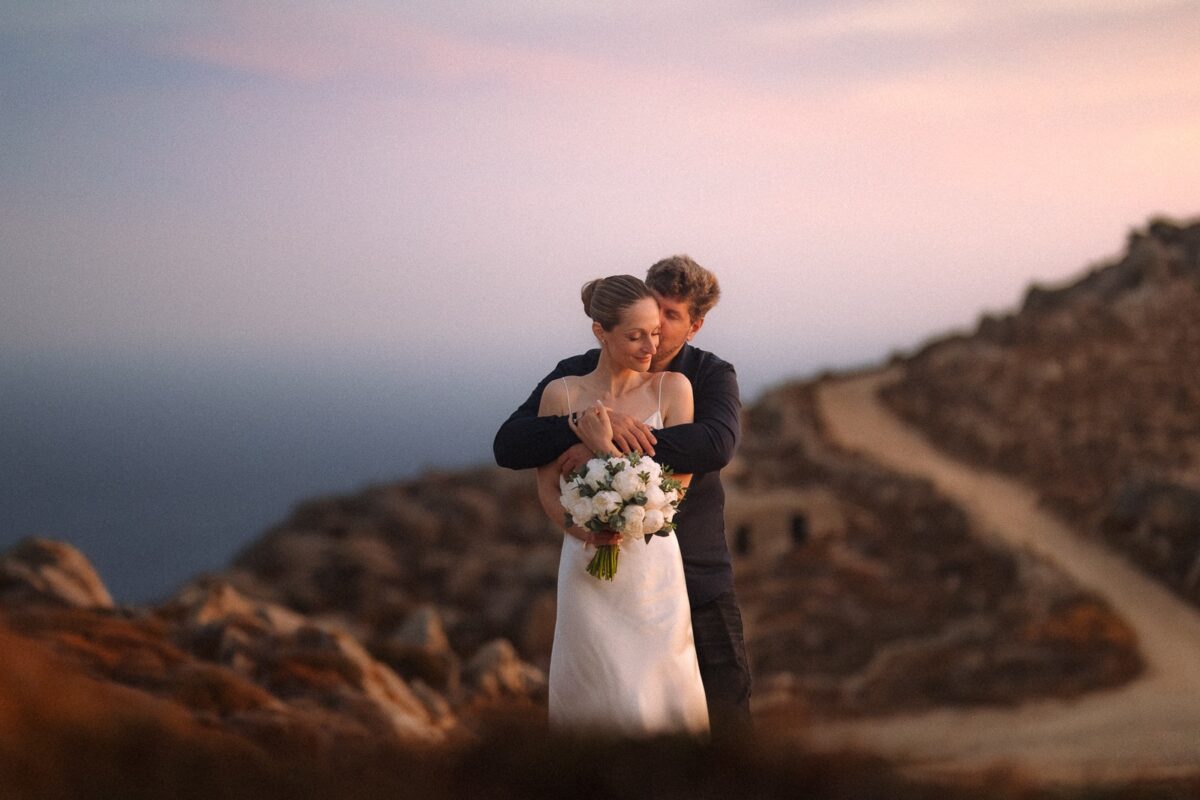Groom holding the bride from behind on a hillside at dusk, with the sea and a winding track behind them