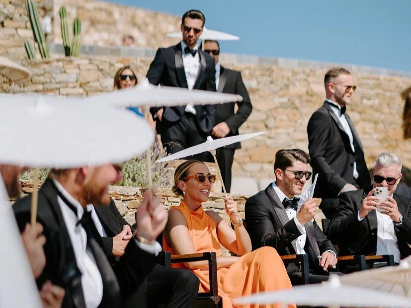 wedding guests dressed in formal wedding attire with gowns and black tie tuxedos sitting under shaded umbrellas at the ceremony