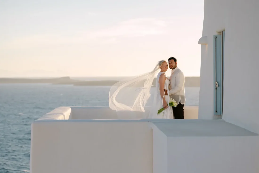 A bride and groom on a whitewashed terrace above the sea on Paros, her veil lifted sideways by the wind