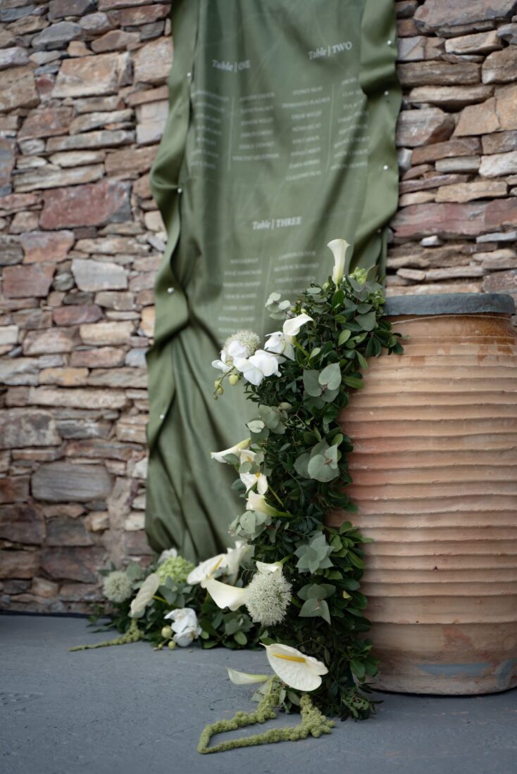 Wedding seating chart on green fabric with white florals against a Sifnos stone wall