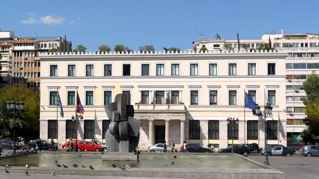 The long neoclassical front of Athens City Hall on Kotzia Square, Greek and European flags flying outside it and a fountain in the foreground. A Greek civil marriage is performed by a mayor or a deputy mayor at a town hall like this one.