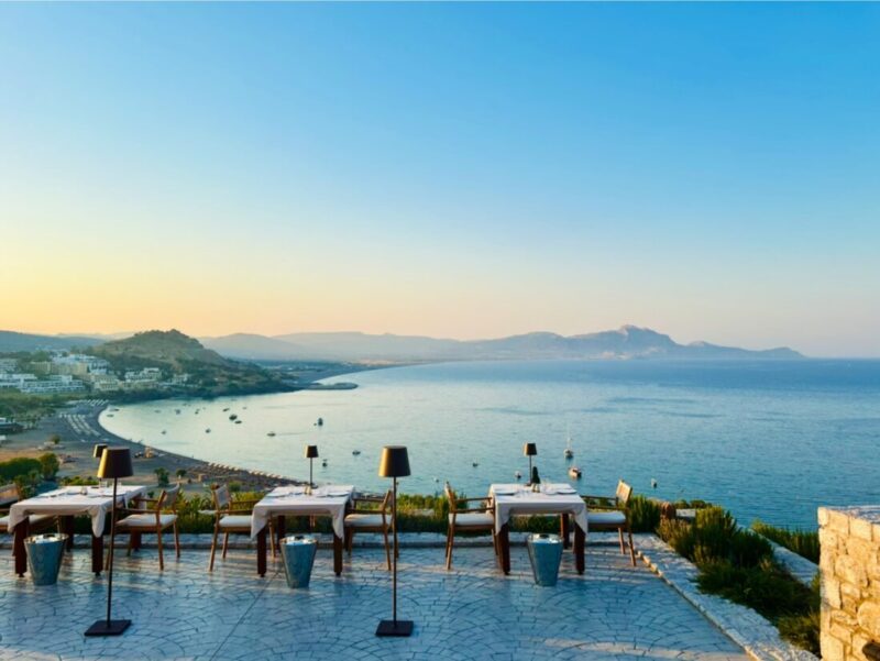 Dining tables along the terrace edge at sunset overlooking Vlicha bay at Ktima Lindos