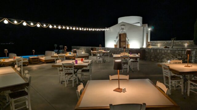 Dining tables lit at night with the chapel floodlit behind at Ktima Lindos