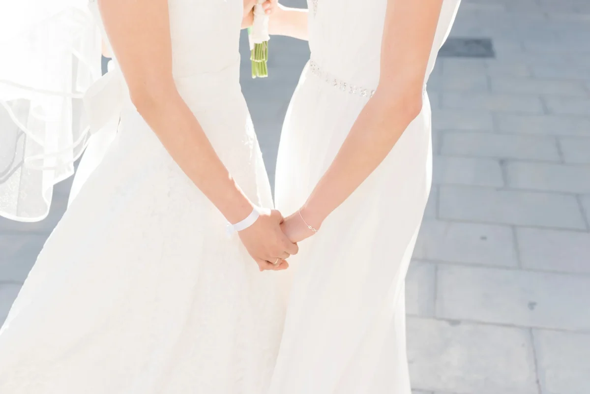Two brides photographed from the shoulders down, hands joined between them, one holding a small white bouquet, standing on sunlit pale stone paving.