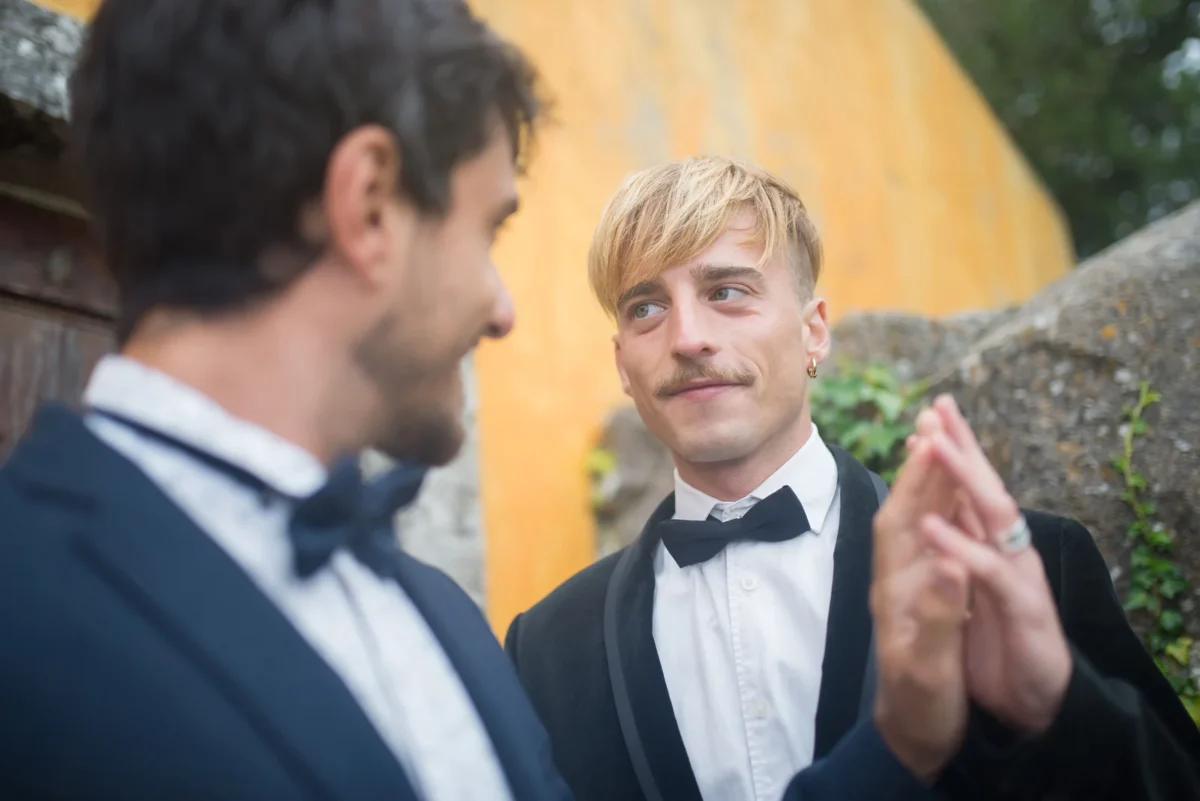 Two grooms in black tie standing close together in front of a weathered ochre wall, one looking at the other, their hands raised and pressed together between them.