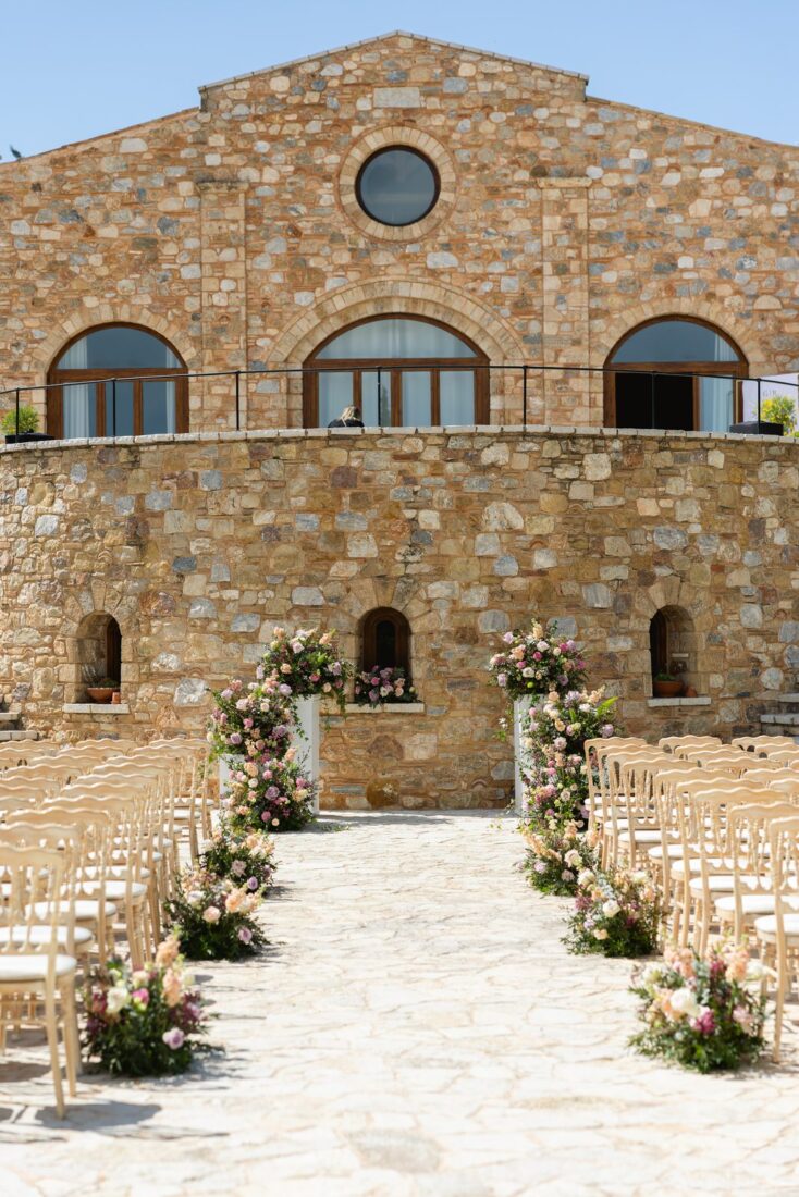 Ceremony aisle lined with pink and white flowers and gold chairs in front of the stone tower at Pyrgos Petreza, a winery near Athens