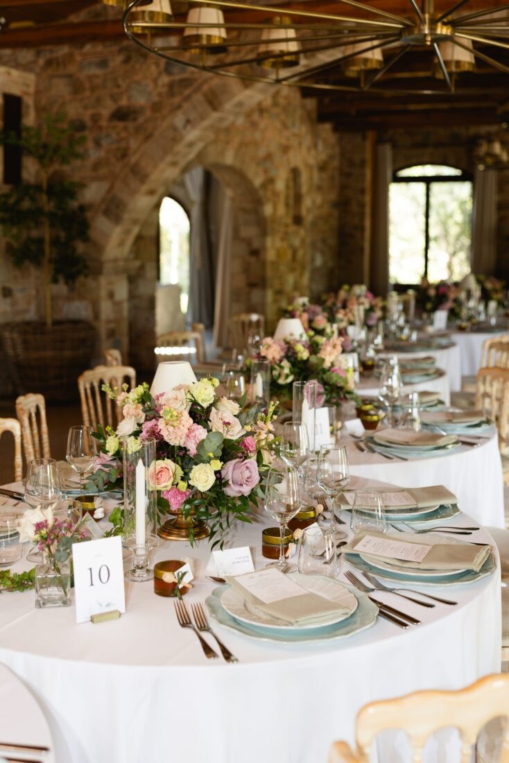 Round wedding reception tables with pink rose centerpieces and sage green plates inside the stone hall at Pyrgos Petreza