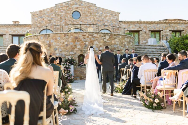 Garanfil walking down the aisle in a long veil between seated guests, toward Roman and the groomsmen, in front of the stone hall at Pyrgos Petreza near Athens