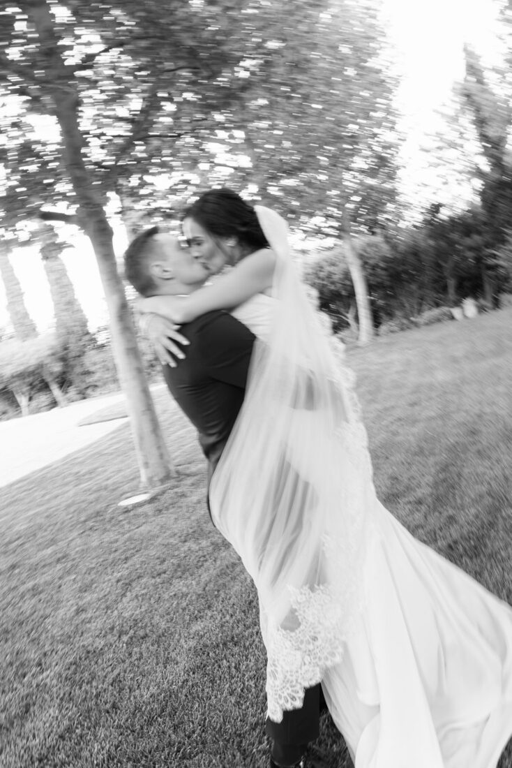 Black and white photo of Roman lifting Garanfil for a kiss under the trees, her veil in motion