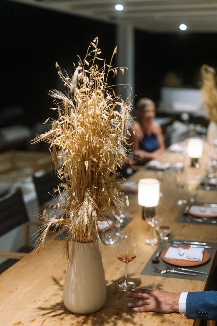 A vase of dried grasses and candle lamps on the wooden dinner table, with family in soft focus behind