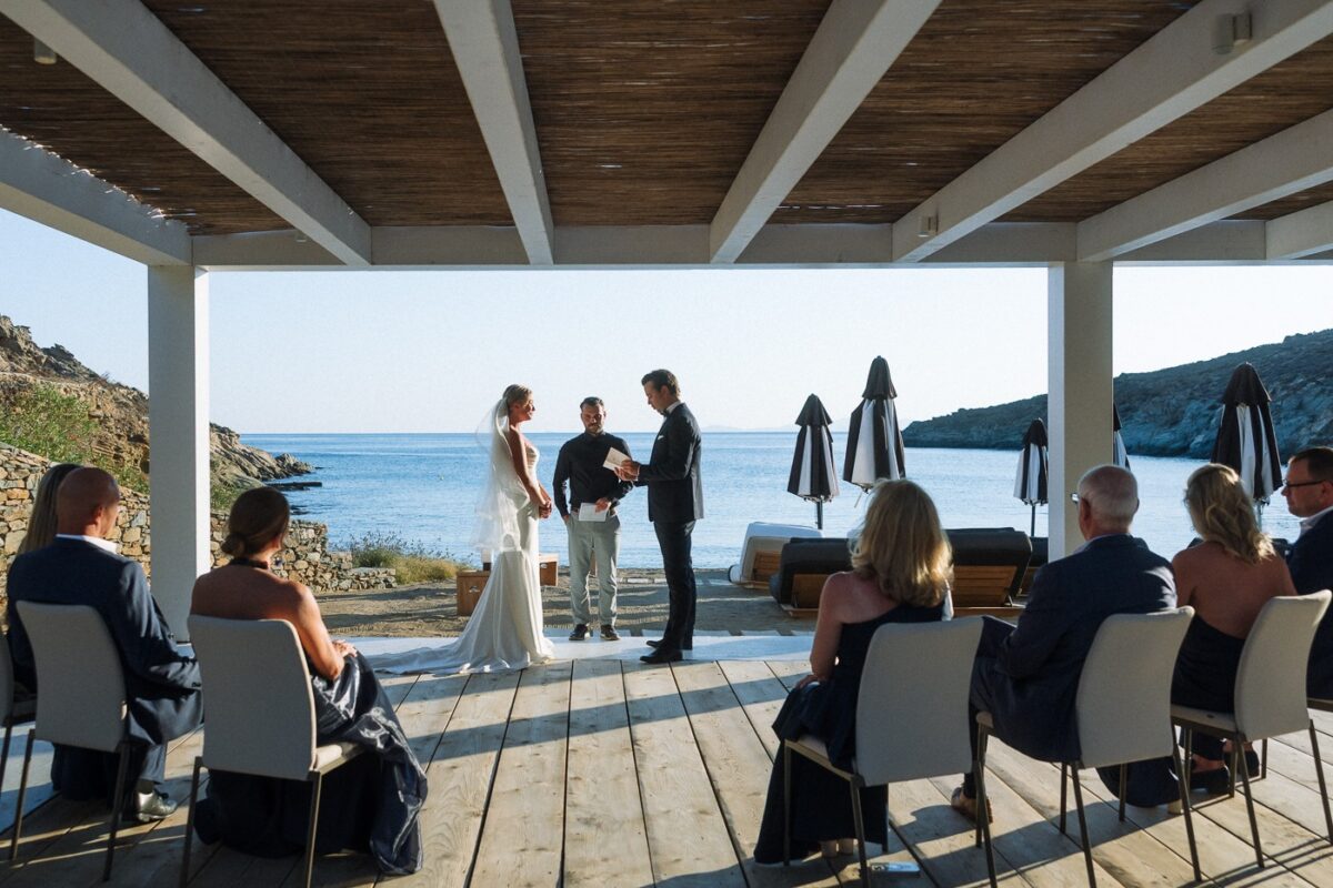 Haley and Mitch's Tinos wedding ceremony on a wooden deck under a white pergola by the sea, with their family seated on both sides
