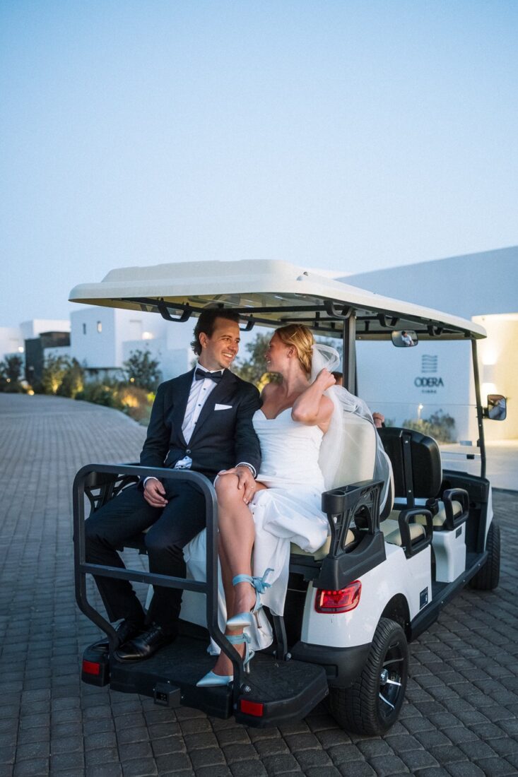 Haley and Mitch laughing on the back of a white golf cart outside the Odera hotel at dusk